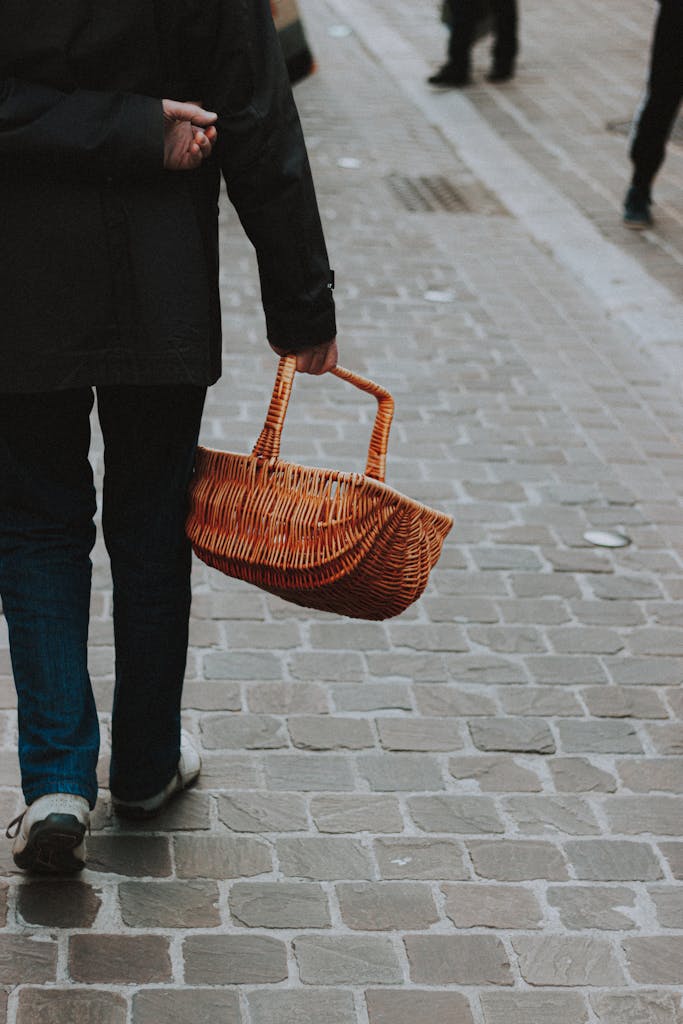 Back view of unrecognizable male in outerwear carrying wicker basket while walking on pavement on street in daytime